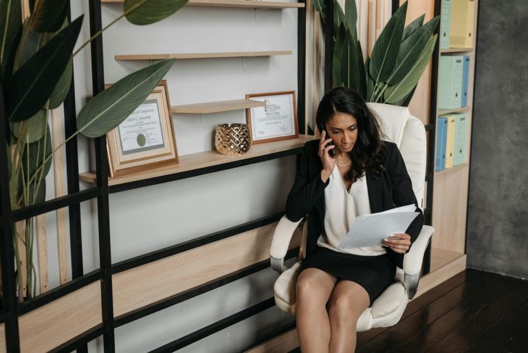 A professional woman in business attire reviews documents while on a phone call in a modern office.